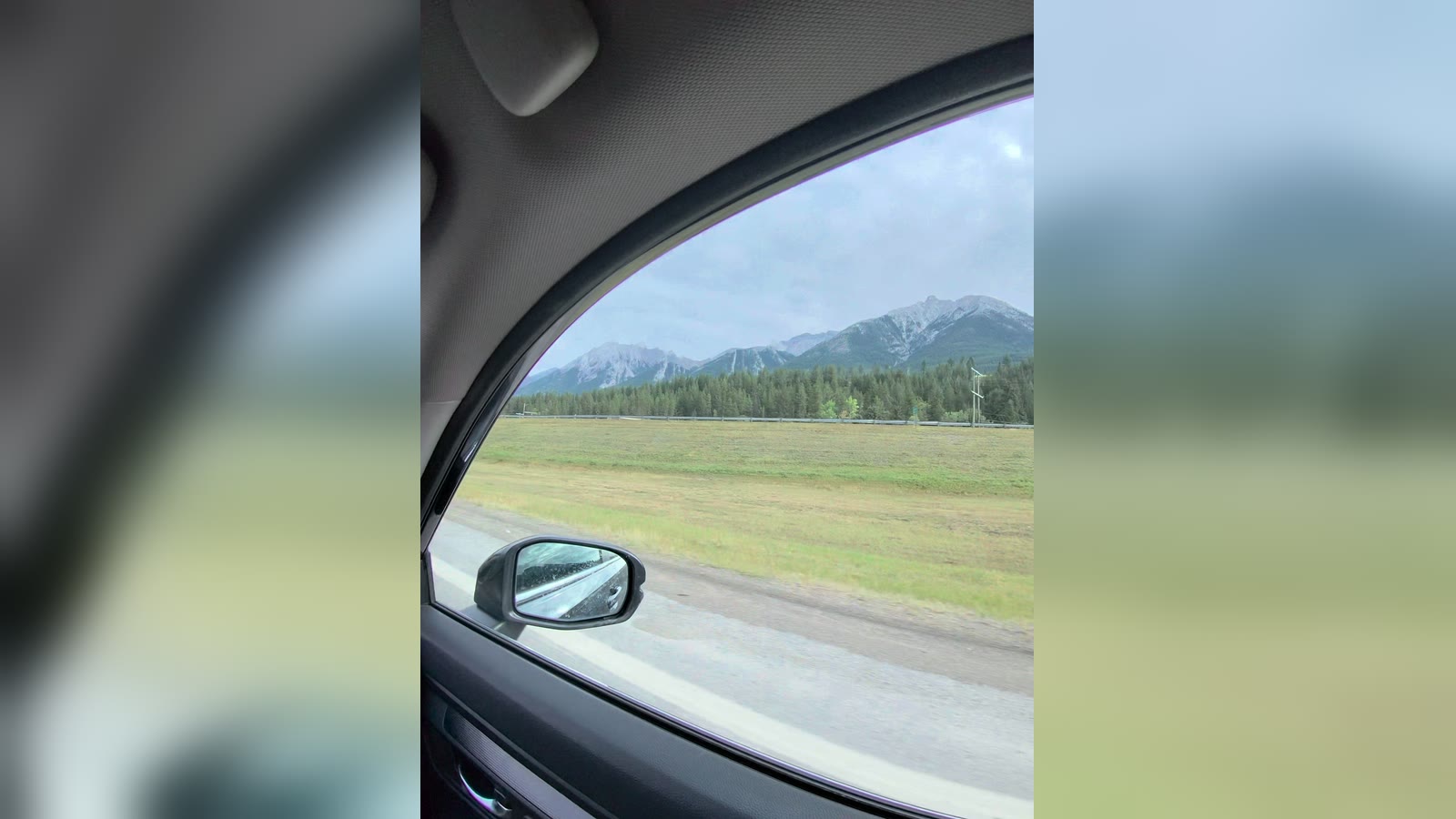 Mountains and pine forest through a car's side window, wing mirror in the corner; an upright clip over a blurred copy of itself